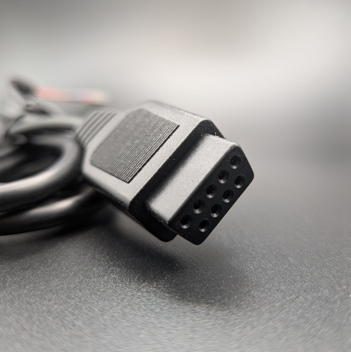 A close-up shot of a black multi-pin electrical connector on a cable, resting on a textured gray surface with a blurred background.