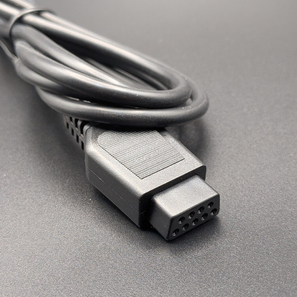 A close-up shot of a black DB9 multi-pin electrical connector on a cable, resting on a textured gray surface with a blurred background.