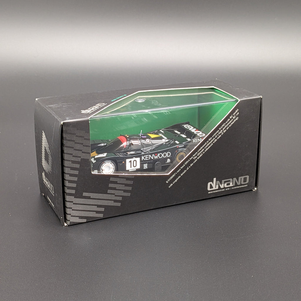 A boxed die-cast model race car displayed in its retail packaging on a dark gray surface. The box is black with a green cutout with a clear window showing a black racing car and visible Kenwood branding.