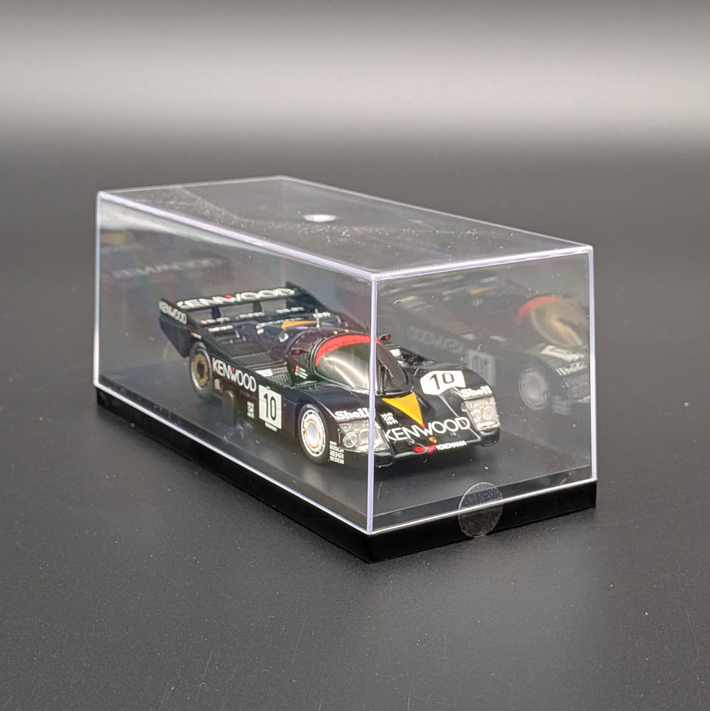 A miniature model of a dark green race car, displayed inside a clear acrylic display case on a black base, set against a dark gray background.