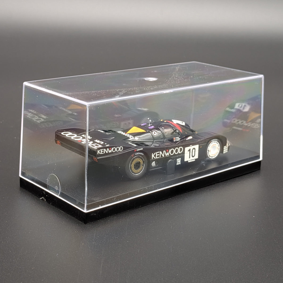 A clear acrylic display case with a black base, housing a black Kenwood race car model. The case is photographed on a dark gray surface against a dark gray background.