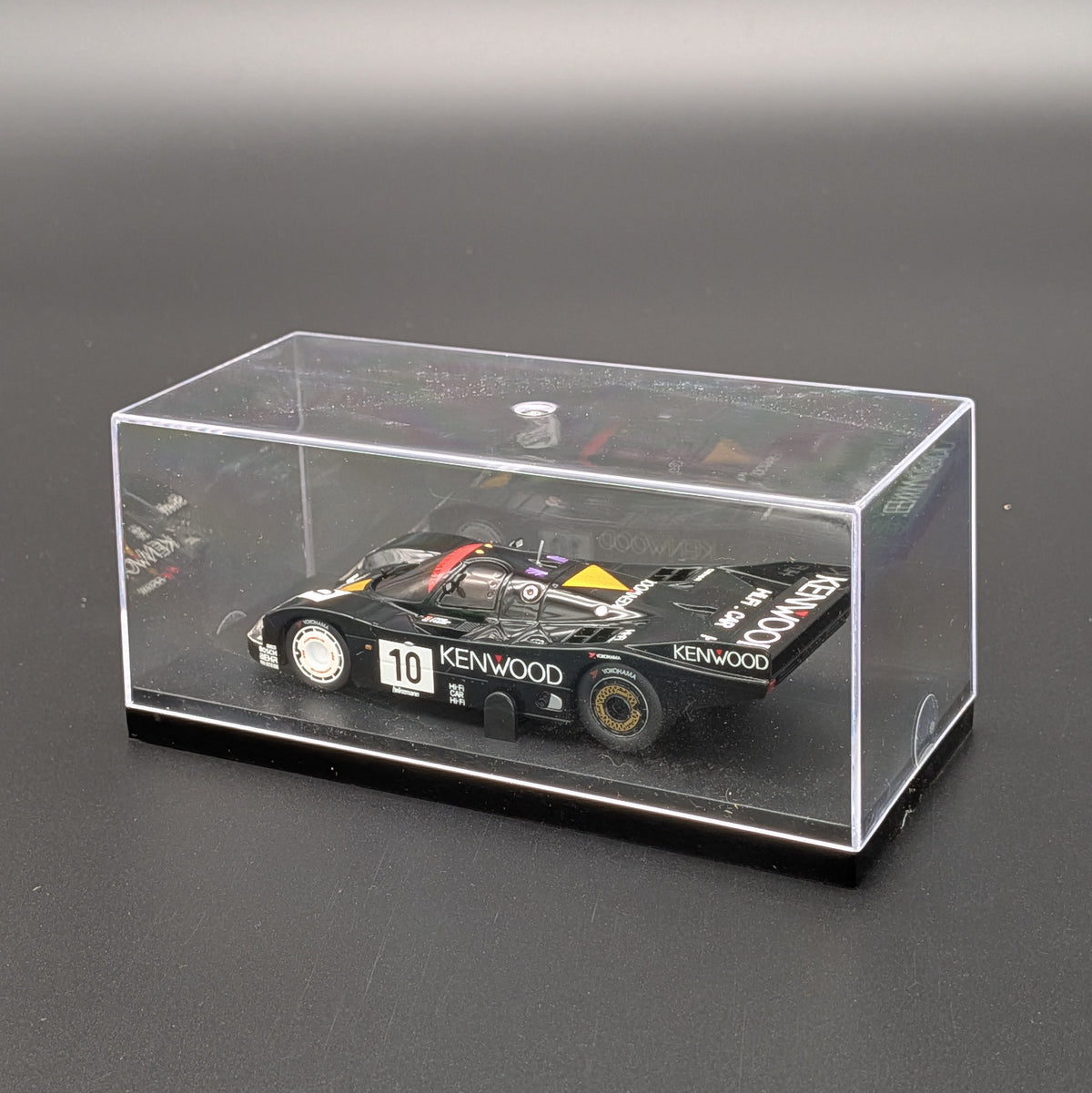 A black Kenwood race car model displayed inside a clear acrylic display case on a dark gray surface.