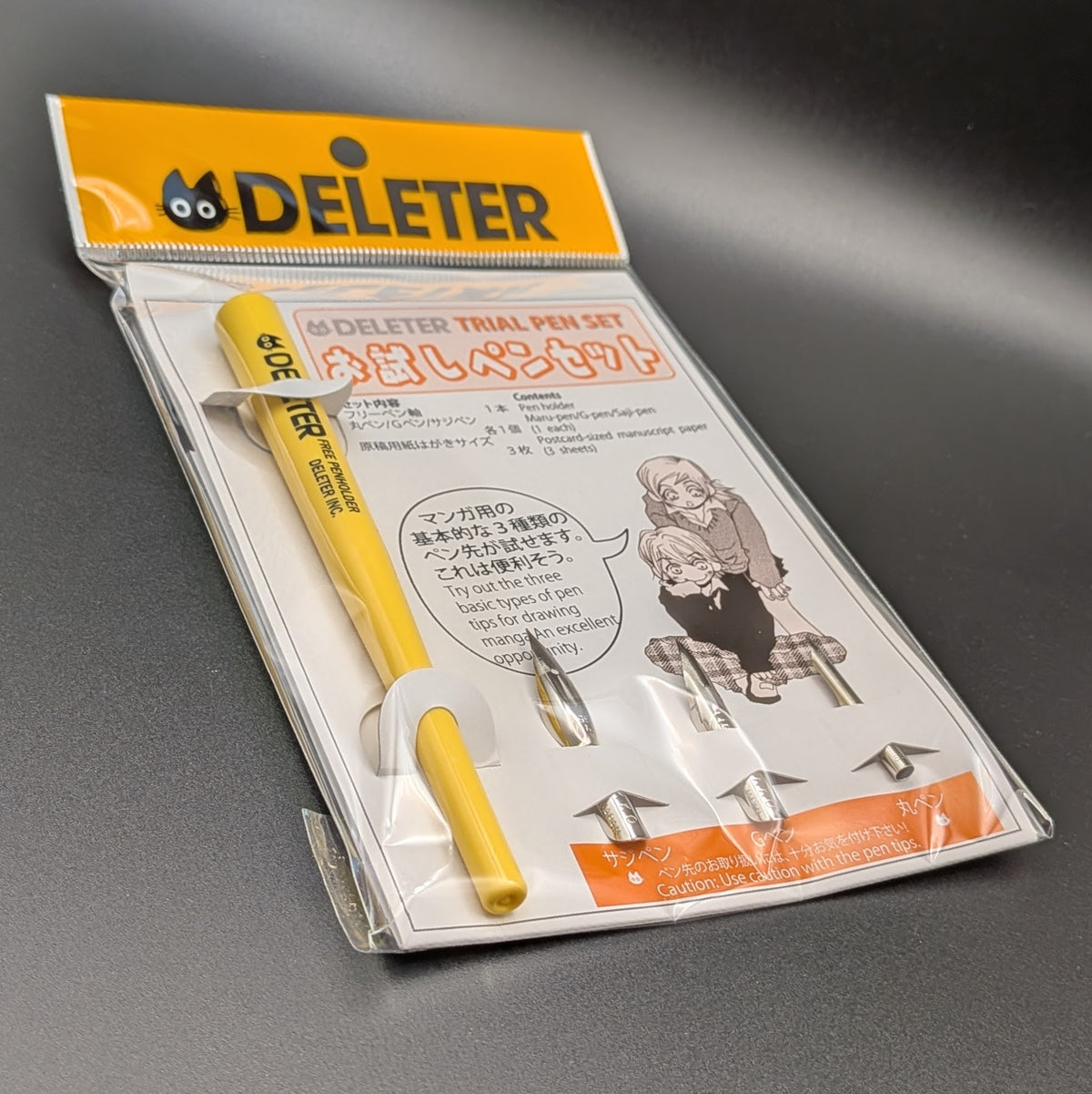 The DELETER Trial Pen Set in its original retail packaging, featuring a yellow pen, 3 pen nibs, manuscript paper, with a white and orange card insert.  The picture shows the item at a 3/4 view angle, leaning to the left, taken on a black background.