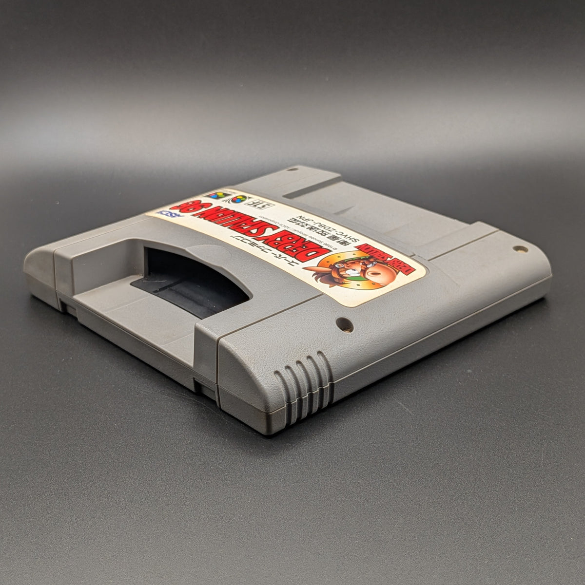 Derby Stallion 96 video game cartridge for Nintendo's Super Famicom (SFC) Entertainment System, shown laying on its back with the label facing upwards. Showing the left hand side and top slot. It sits on a dark grey background.