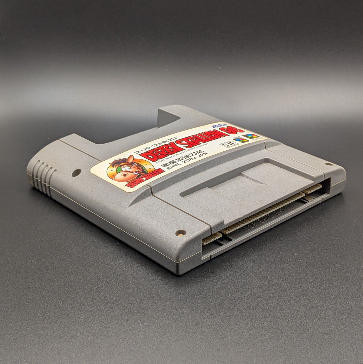 Derby Stallion 96 video game cartridge for Nintendo's Super Famicom (SFC) Entertainment System, shown laying on its back with the label facing upwards. Showing the left hand side and bottom slot. It sits on a dark grey background.