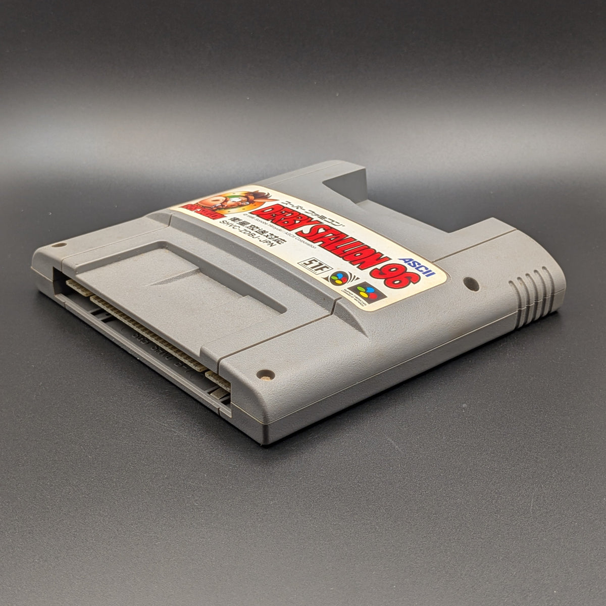 Derby Stallion 96 video game cartridge for Nintendo's Super Famicom (SFC) Entertainment System, shown laying on its back with the label facing upwards. Showing the right hand side and bottom slot. It sits on a dark grey background.