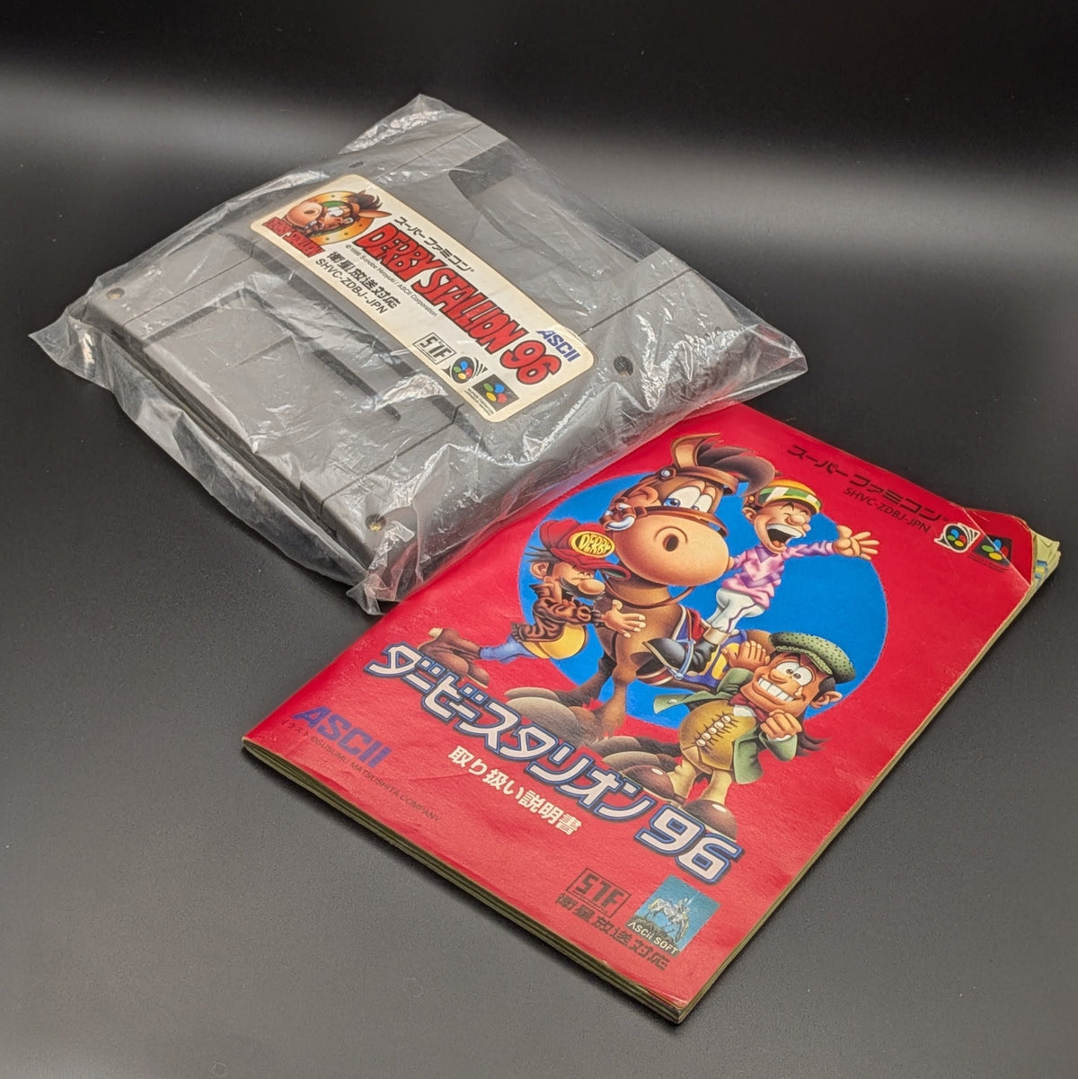 Derby Stallion 96 video game cartridge in plastic for Nintendo's Super Famicom (SFC) Entertainment System, shown laying on its back, label facing upwards next to its manual. The manual has a crease in the top right corner. They are displayed on a dark grey background.