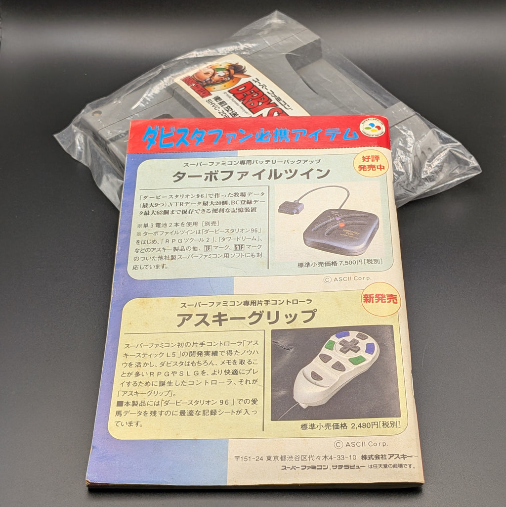 Derby Stallion 96 video game cartridge in plastic for Nintendo's Super Famicom (SFC) Entertainment System, shown laying on its back, label facing upwards with its manual half laid over the top of the cart and shows the back. The back of the manual depicts adverts for console accessories in Japanese. They are displayed on a dark grey background.