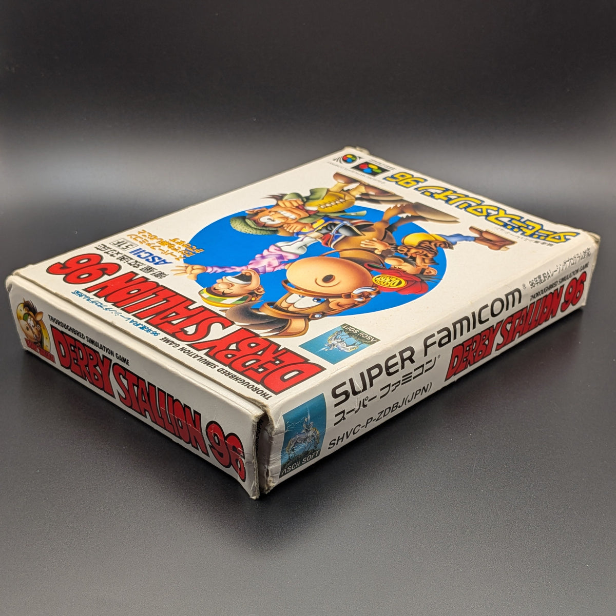 Derby Stallion 96 boxed video game for Nintendo's Super Famicom (SFC) Entertainment System, shown laying on its back with the front cover facing upwards. Showing the left hand side and top edge. It sits on a dark grey background. There is some wear and tear on the top left corner, most likely due to age.