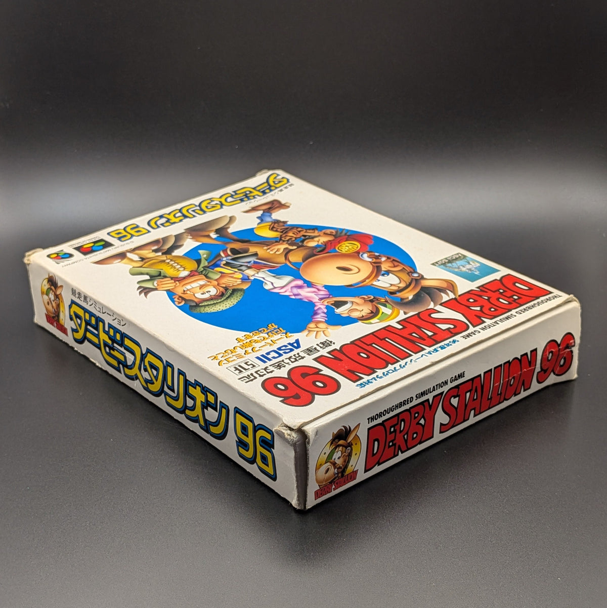 Derby Stallion 96 boxed video game for Nintendo's Super Famicom (SFC) Entertainment System, shown laying on its back with the front cover facing upwards. Showing the right hand side and top edge. It sits on a dark grey background. There is some wear and tear on the top right corner, most likely due to age.