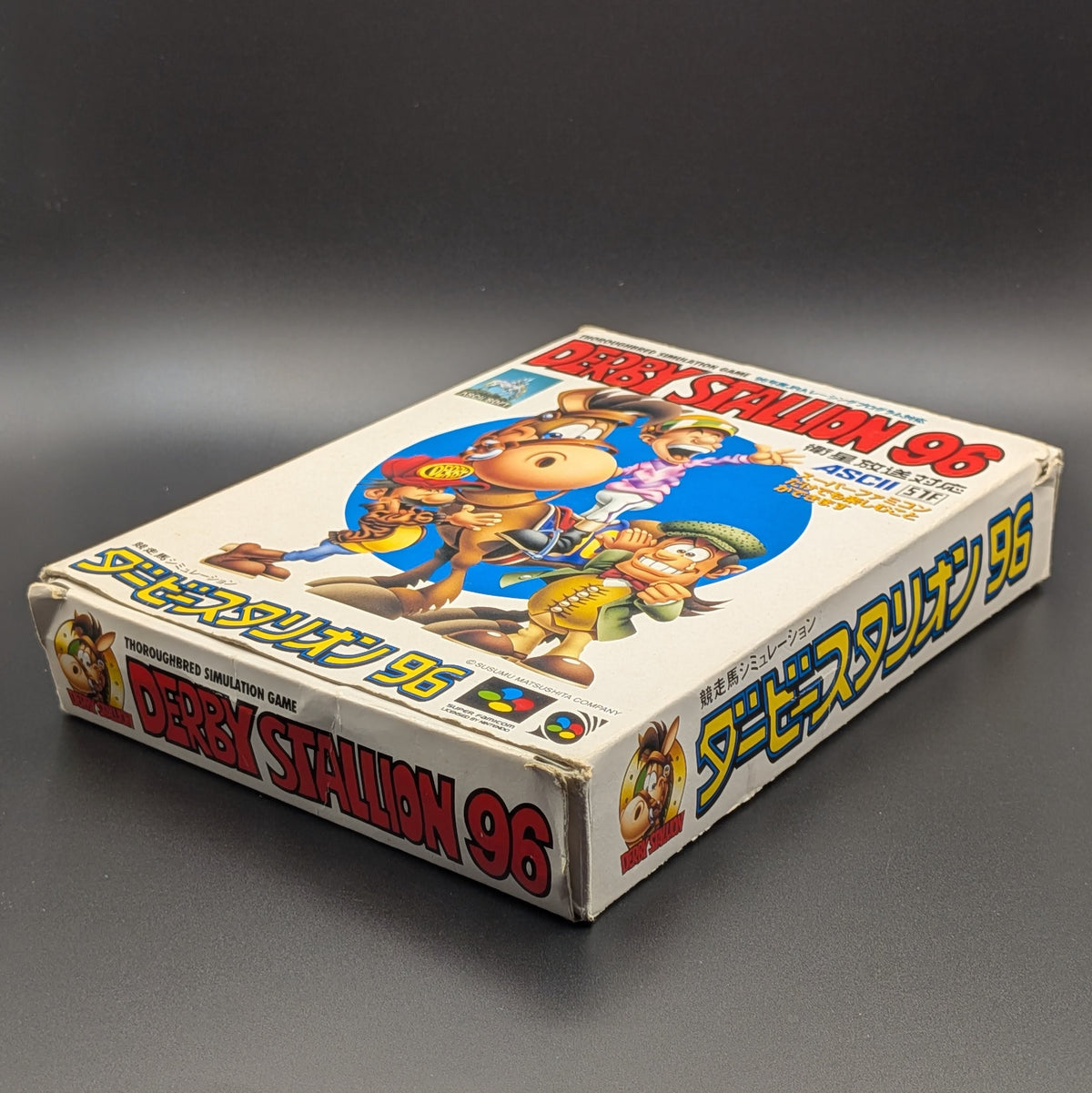 Derby Stallion 96 boxed video game for Nintendo's Super Famicom (SFC) Entertainment System, shown laying on its back with the front cover facing upwards. Showing the right hand side and bottom edge. It sits on a dark grey background. There is some wear and tear on the bottom corners, most likely due to age.