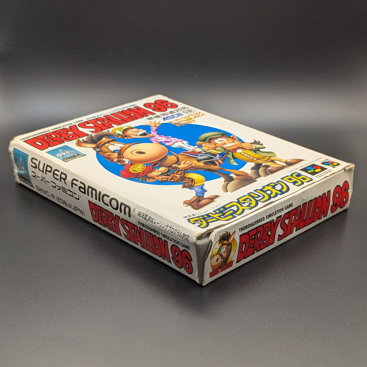 Derby Stallion 96 boxed video game for Nintendo's Super Famicom (SFC) Entertainment System, shown laying on its back with the front cover facing upwards. Showing the left hand side and bottom edge. It sits on a dark grey background. There is some wear and tear on the bottom left corner, most likely due to age.