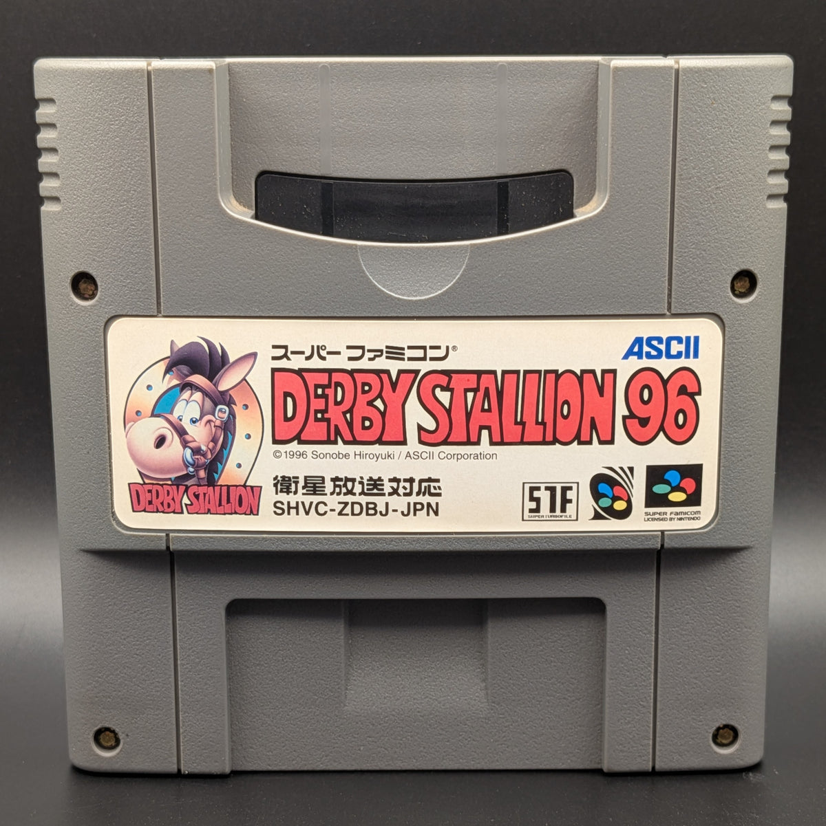 A close up of Derby Stallion 96' video game cartridge for Nintendo's Super Famicom (SFC) Entertainment System, shown standing up and facing forward on a dark grey background.
