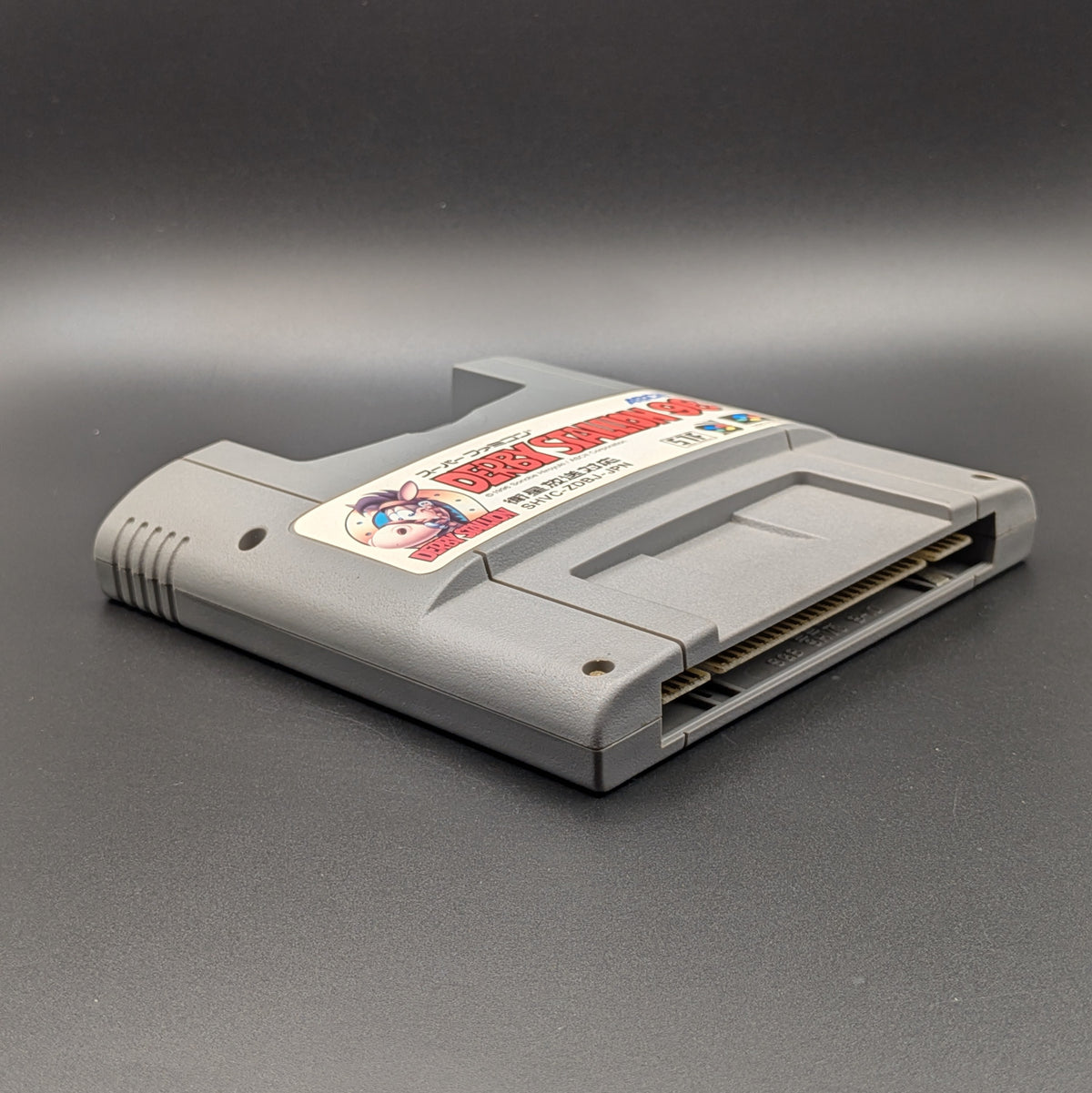 Derby Stallion 96 video game cartridge for Nintendo's Super Famicom (SFC) Entertainment System, shown laying on its back with the label facing upwards. Showing the left hand side and bottom slot. It sits on a dark grey background.