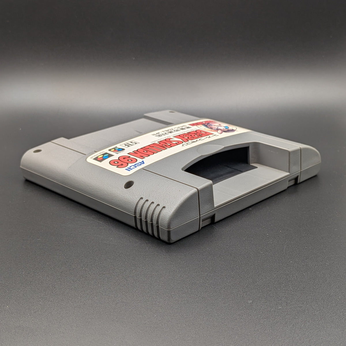 Derby Stallion 96 video game cartridge for Nintendo's Super Famicom (SFC) Entertainment System, shown laying on its back with the label facing upwards. Showing the right hand side and top section. It sits on a dark grey background.
