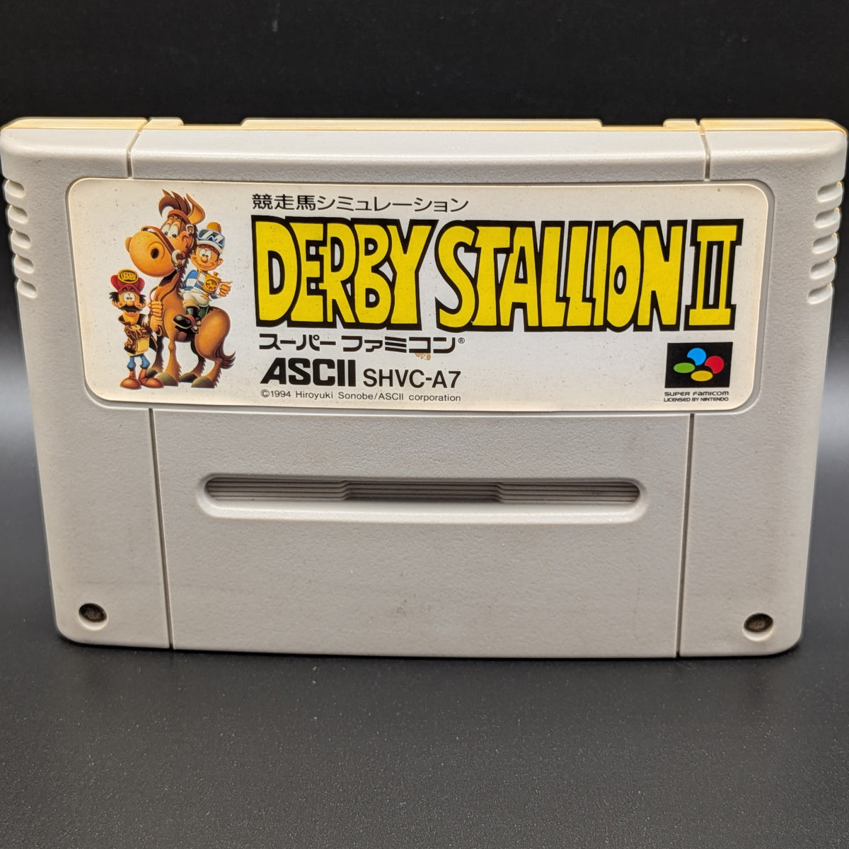 A close up of Derby Stallion 2 video game cartridge for Nintendo's Super Famicom (SFC) Entertainment System, shown standing up and facing forwards on a dark grey background.

