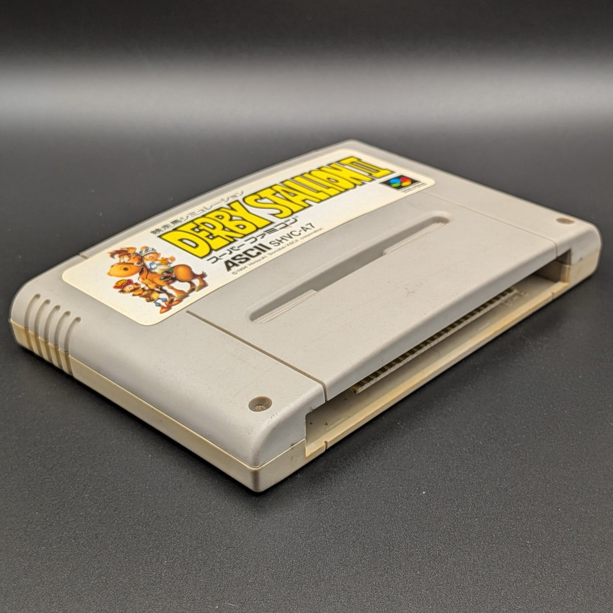 Derby Stallion 2  video game cartridge for Nintendo's Super Famicom (SFC) Entertainment System, shown laying on its back with the label facing upwards. Showing the left hand side and bottom slot. It sits on a dark grey background.