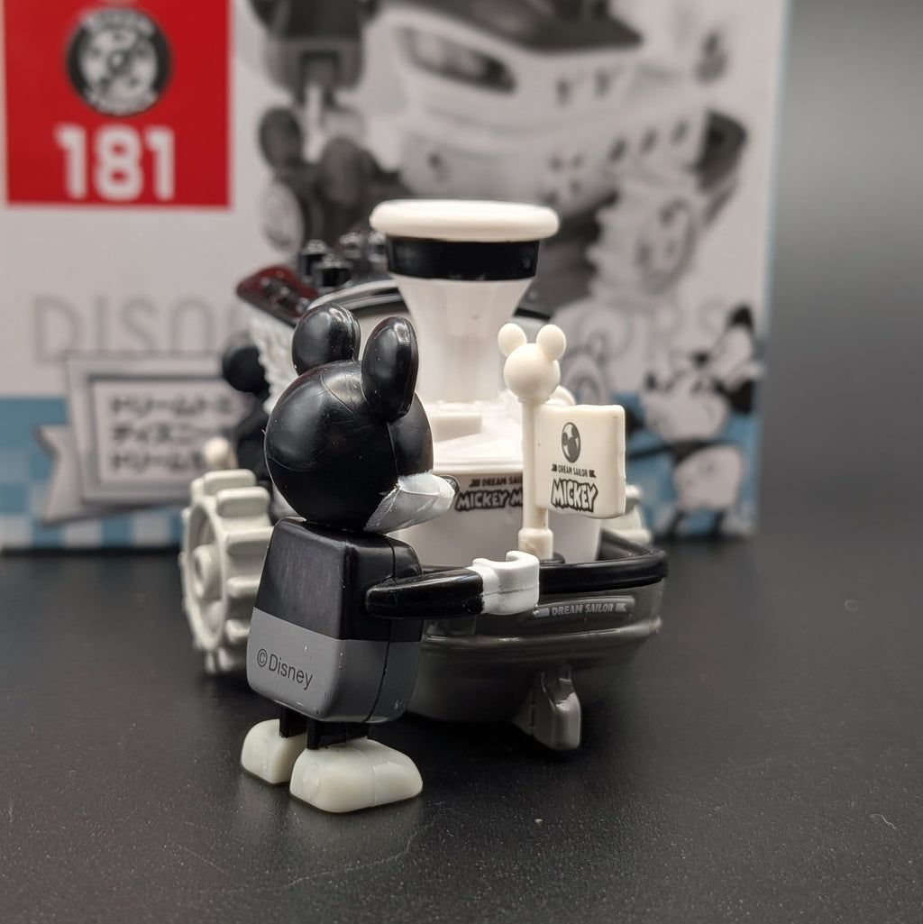 A Disney Mickey Mouse toy figure in a black and white outfit standing next to a boat, photographed on a dark surface with the product box blurred in the background.