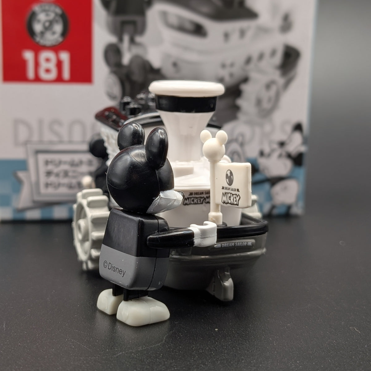A Disney Mickey Mouse toy figure in a black and white outfit standing next to a boat, photographed on a dark surface with the product box blurred in the background.