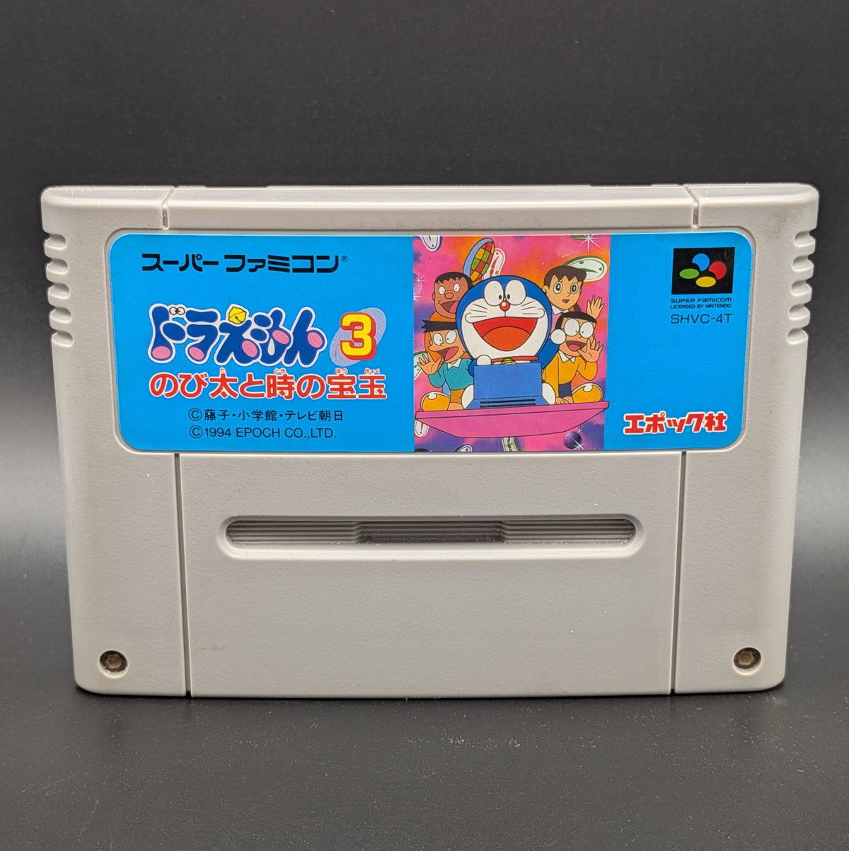 A close up of Doraemon 3 video game cartridge for Nintendo's Super Famicom (SFC) Entertainment System, shown standing up and facing forwards on a dark grey background.