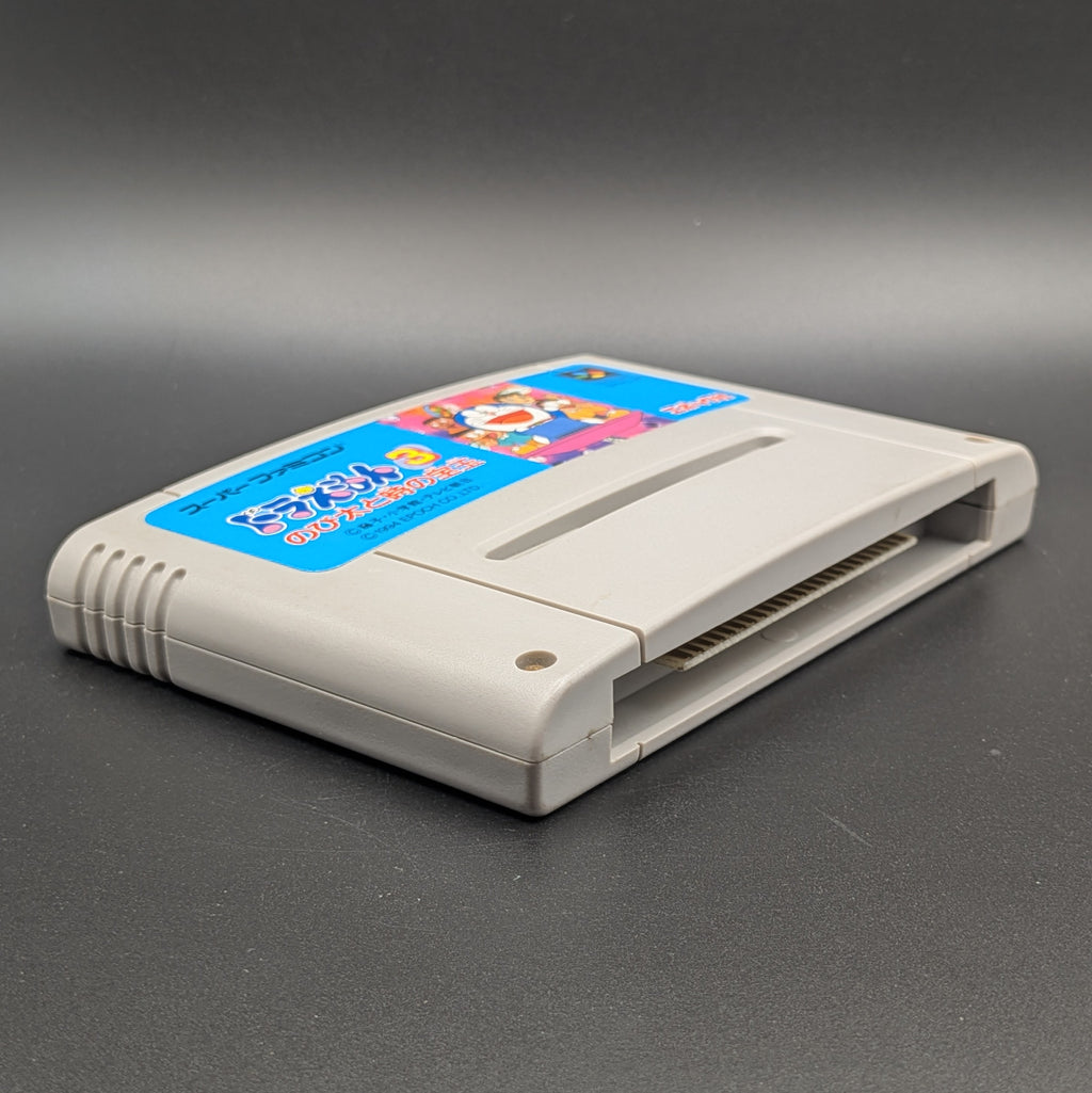 Doraemon 3 video game cartridge for Nintendo's Super Famicom (SFC) Entertainment System, shown laying on its back with the label facing upwards. Showing the left hand side and bottom slot. It sits on a dark grey background. 
