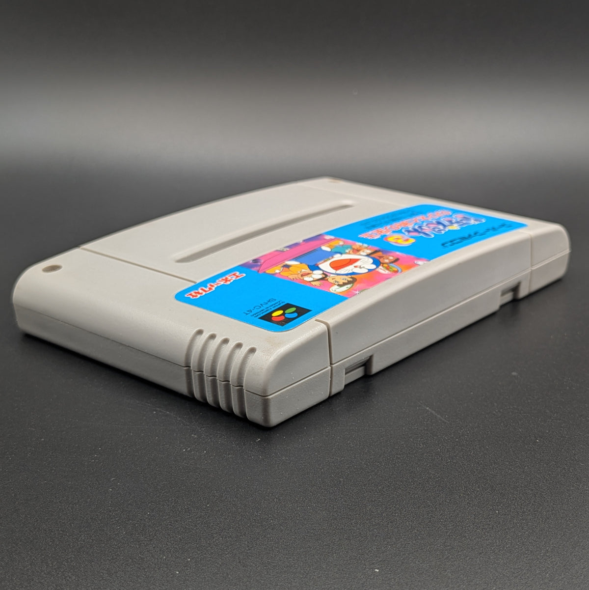 Doraemon 3 video game cartridge for Nintendo's Super Famicom (SFC) Entertainment System, shown laying on its back with the label facing upwards. Showing the right hand side and top edge. It sits on a dark grey background. 