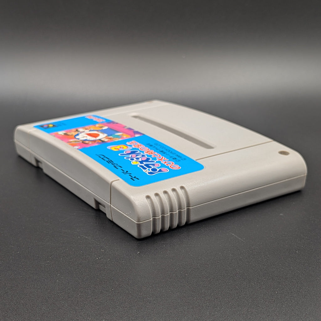 Doraemon 3 video game cartridge for Nintendo's Super Famicom (SFC) Entertainment System, shown laying on its back with the label facing upwards. Showing the left hand side and top edge. It sits on a dark grey background. 