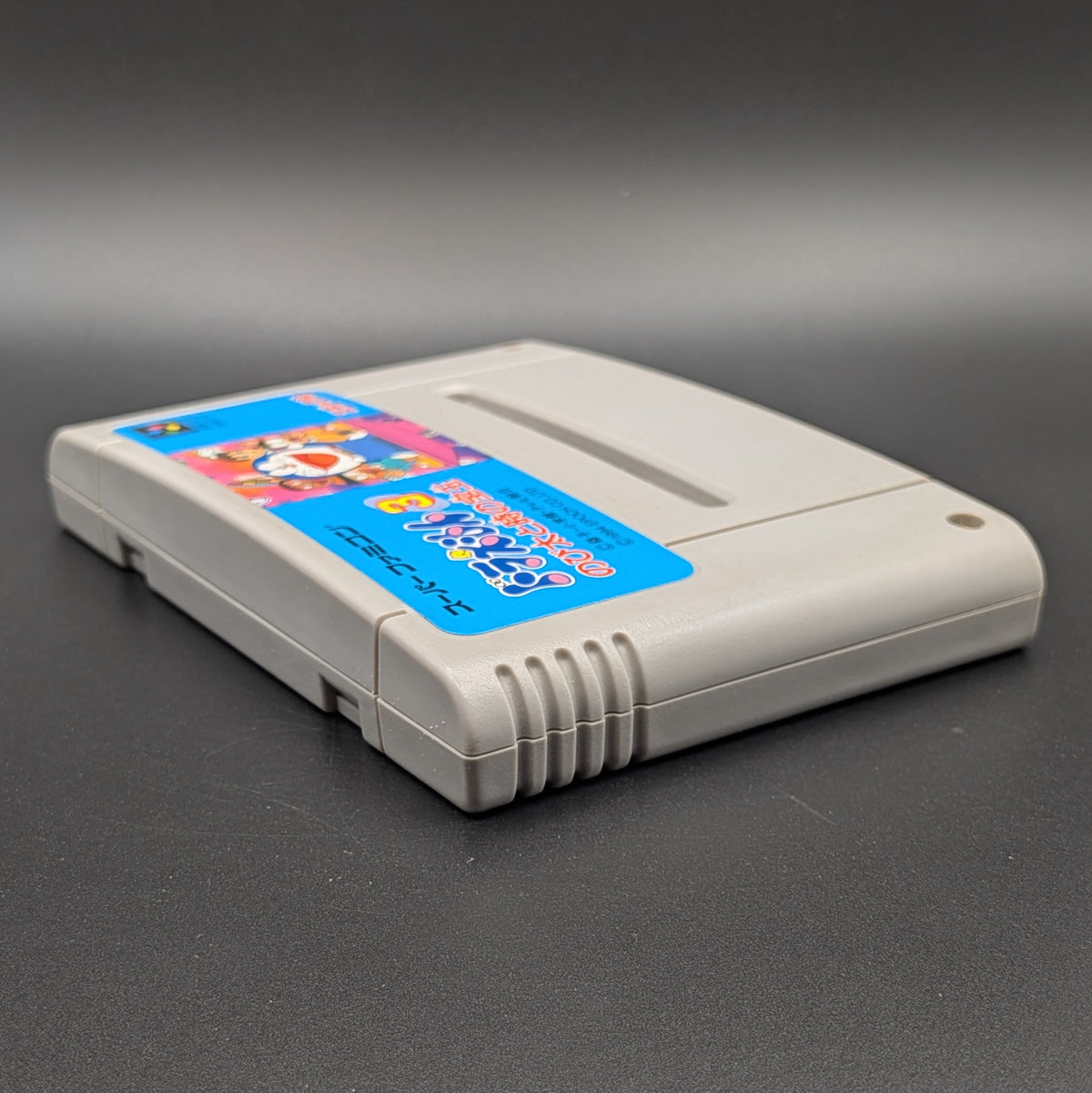 Doraemon 3 video game cartridge for Nintendo's Super Famicom (SFC) Entertainment System, shown laying on its back with the label facing upwards. Showing the left hand side and top edge. It sits on a dark grey background. 