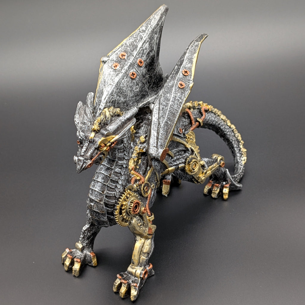 A steampunk-style fantasy dragon figurine with metallic silver scales, gold gears, and copper accents, standing on a dark gray background.