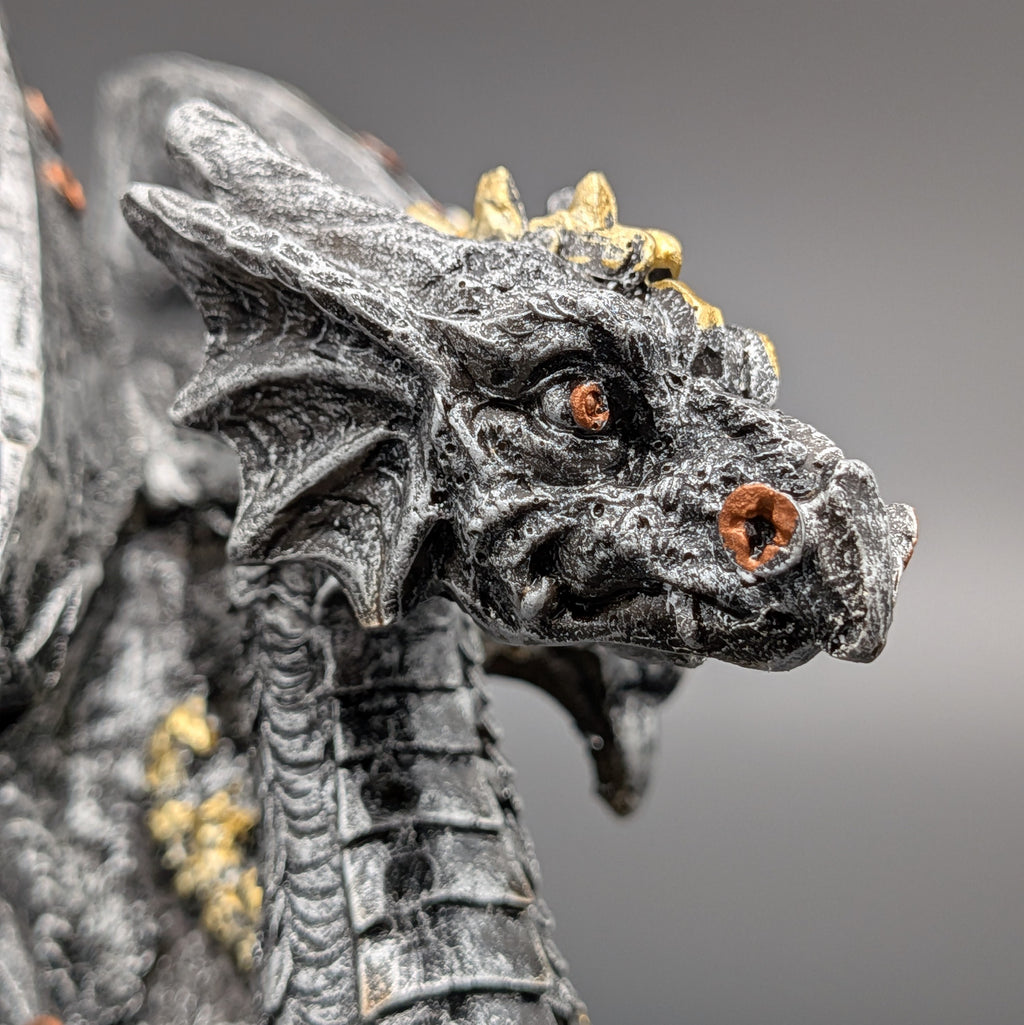 Close-up of a detailed fantasy dragon figurine with textured dark gray scales, small gold horns, and orange eyes against a soft gray background.