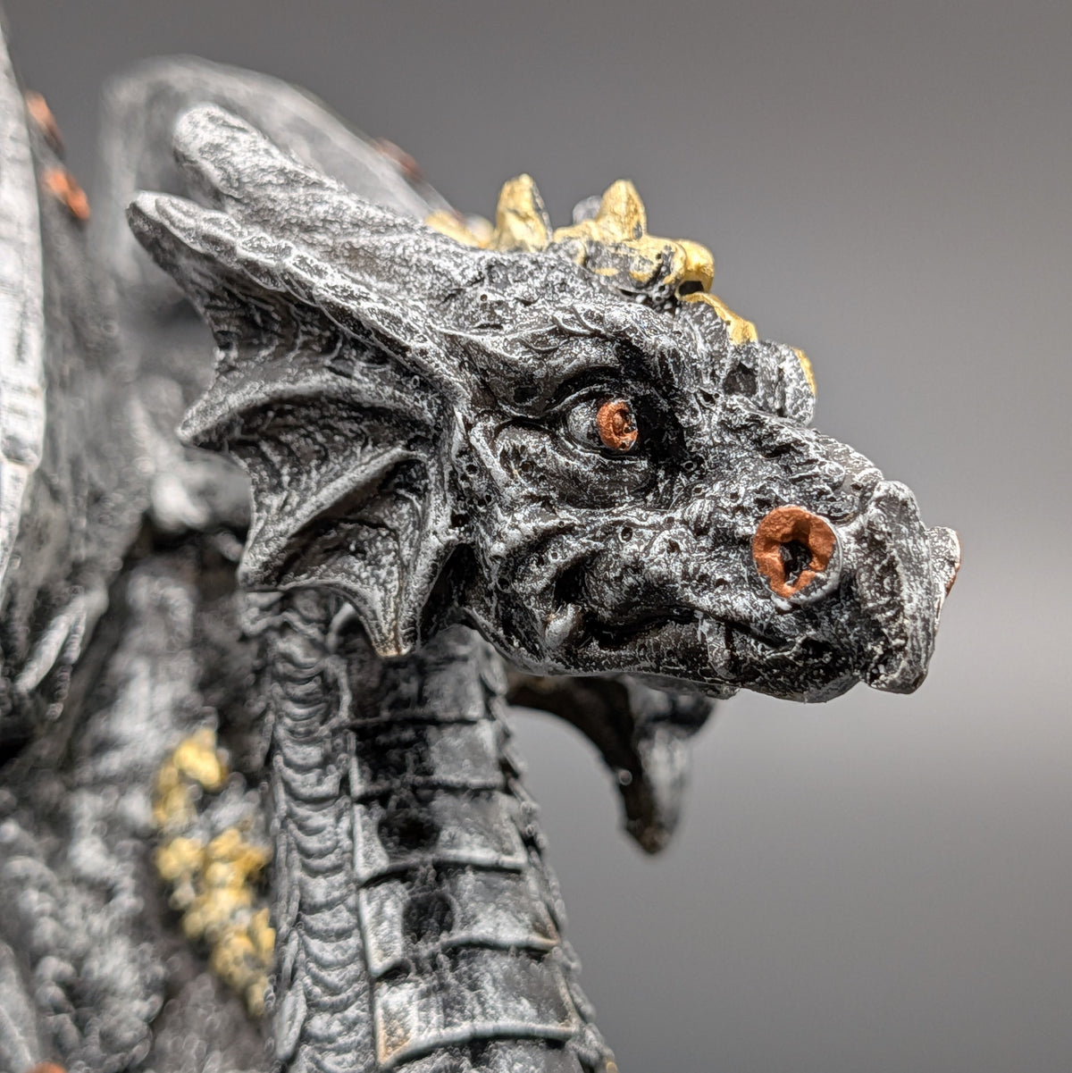 Close-up of a detailed fantasy dragon figurine with textured dark gray scales, small gold horns, and orange eyes against a soft gray background.