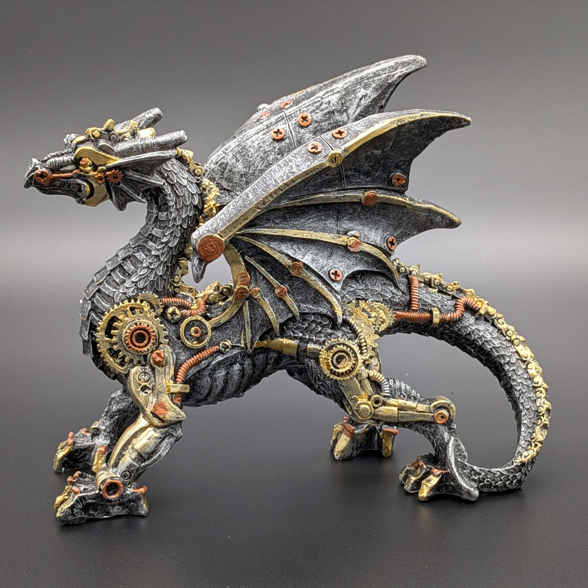 A steampunk-style fantasy dragon figurine with metallic gray scales, gold gears, and copper mechanical details, standing on a dark gray surface against a gradient gray background.