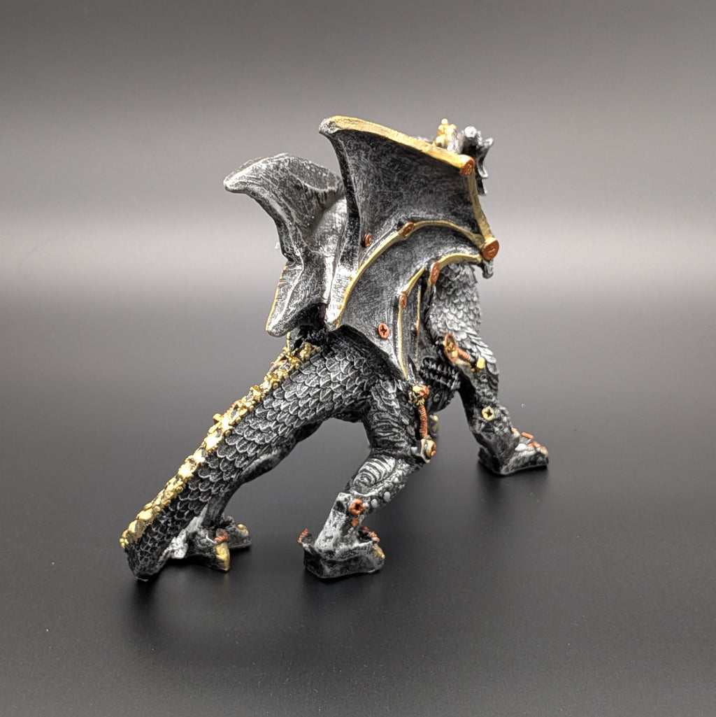 A fantasy dragon figurine standing on a dark gray surface against a matching gray gradient background. The dragon is primarily textured black with metallic gold accents along its wings, tail, and joints.