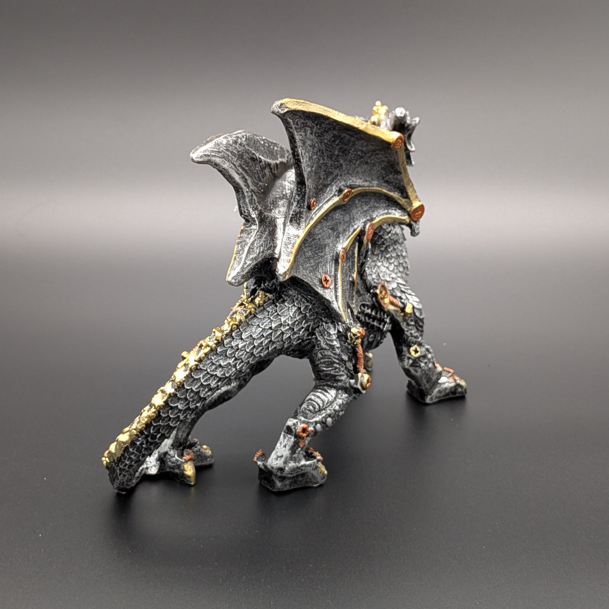 A fantasy dragon figurine standing on a dark gray surface against a matching gray gradient background. The dragon is primarily textured black with metallic gold accents along its wings, tail, and joints.