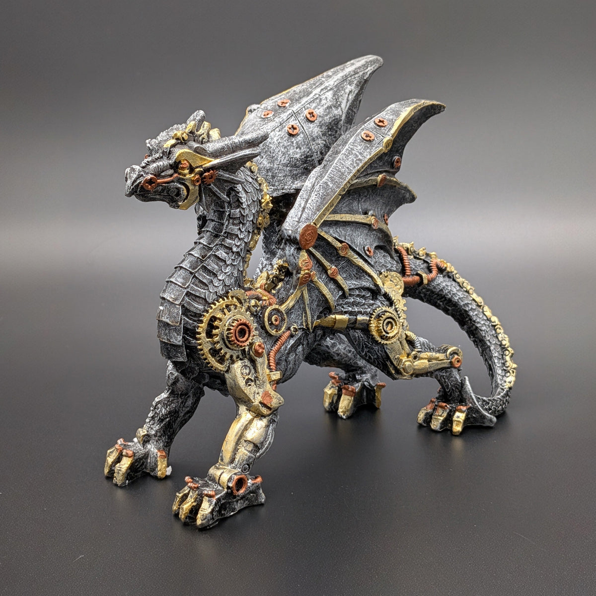 A steampunk-style fantasy dragon figurine with metallic gray scales, gold gears, and copper-toned mechanical details, photographed on a dark gray studio background.