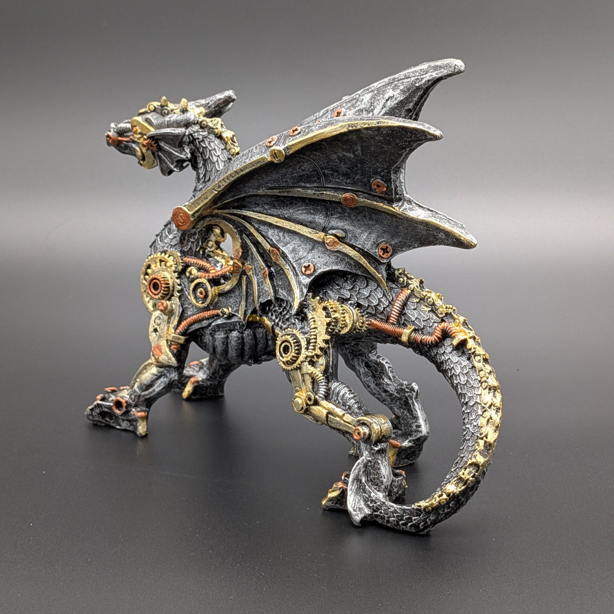 Steampunk dragon figurine with intricate metal gears, cogs, and pipes, featuring a dark metallic gray body and wings with gold and copper accents, set against a dark gray gradient background.