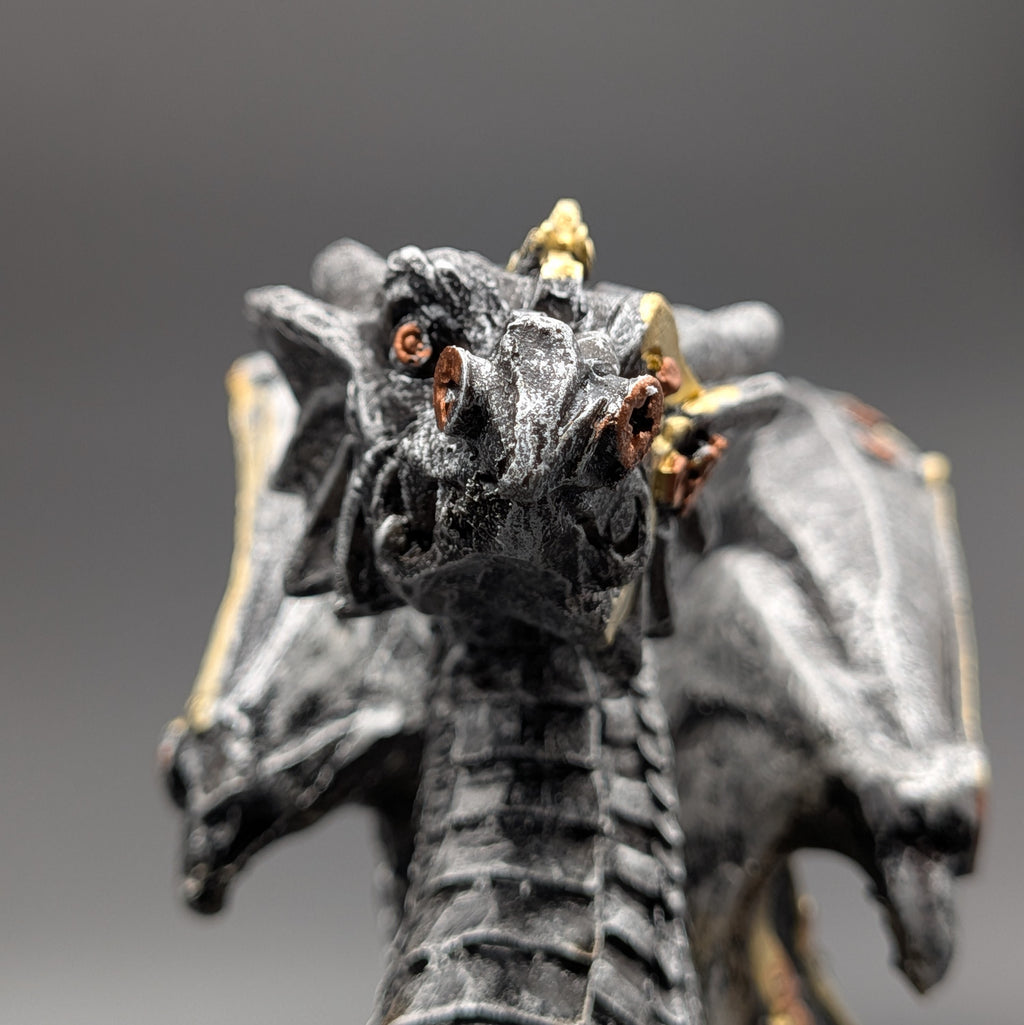 A close-up of a detailed fantasy dragon figurine with a textured black and gray body, gold accents on its horns and wings, and small orange eyes, set against a dark gray gradient background.