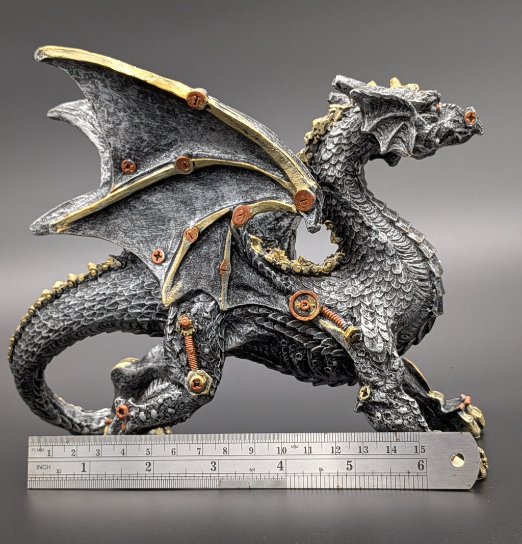 A steampunk-style dragon figurine with a textured dark gray body, gold-toned mechanical accents, and visible screws and springs, shown with a metal ruler at the bottom for scale against a dark gray gradient background.