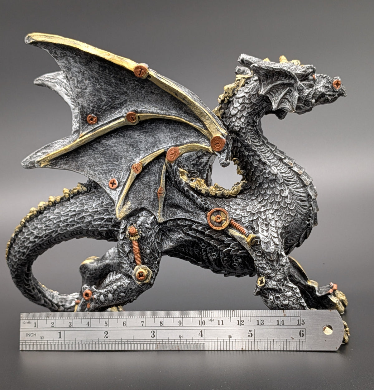 A steampunk-style dragon figurine with a textured dark gray body, gold-toned mechanical accents, and visible screws and springs, shown with a metal ruler at the bottom for scale against a dark gray gradient background.