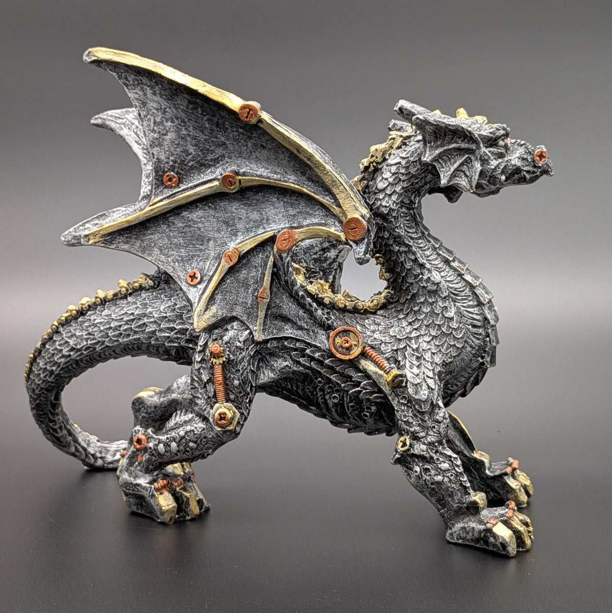 A detailed fantasy dragon figurine in a dynamic pose, featuring a textured dark gray and black scale pattern with metallic gold accents on its wings, claws, and joints, set against a dark gray gradient background.