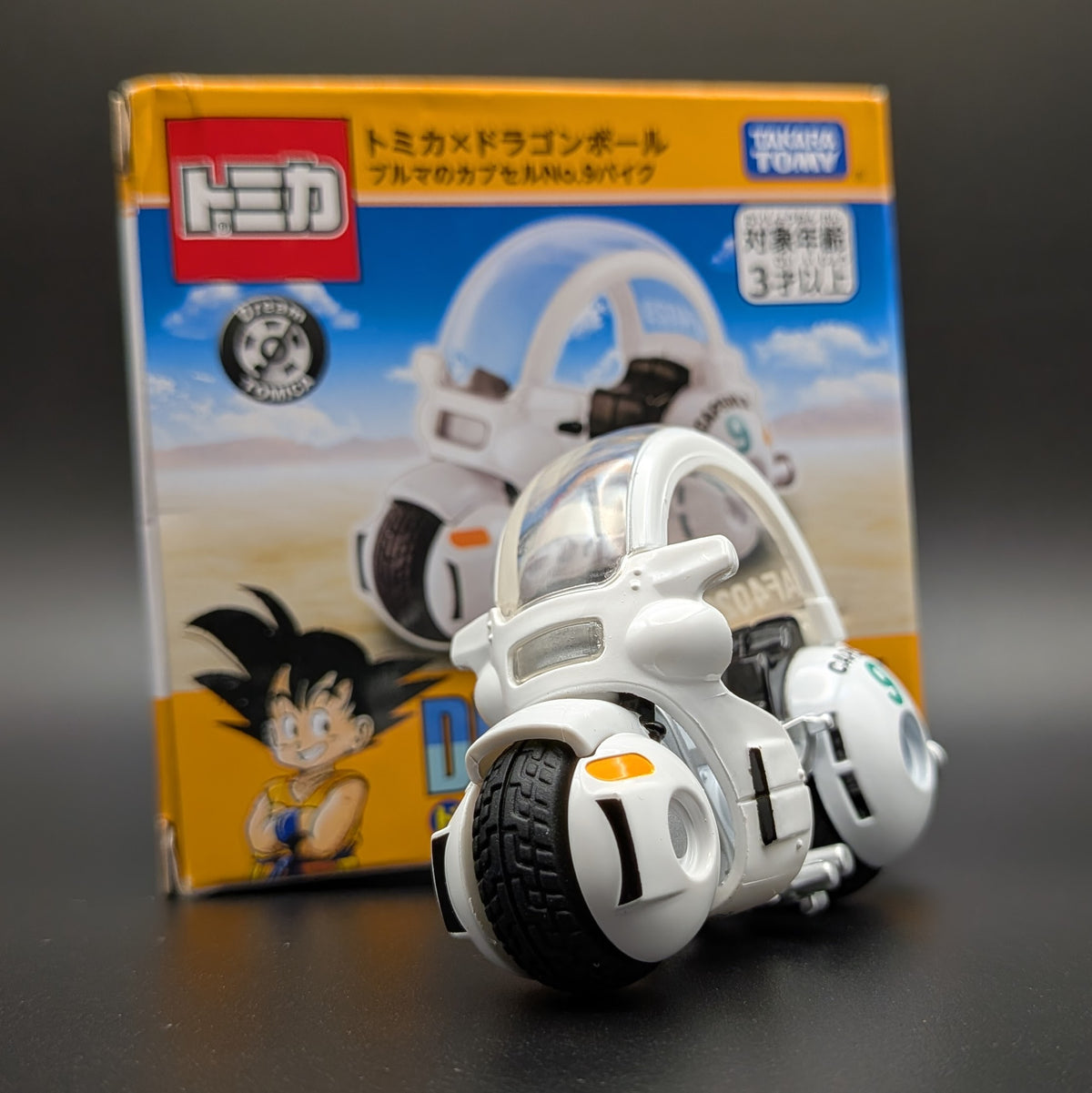 A white and black Tomica Dragon Ball toy motorcycle with a clear canopy, displayed in front of its original yellow and blue packaging box on a dark surface.