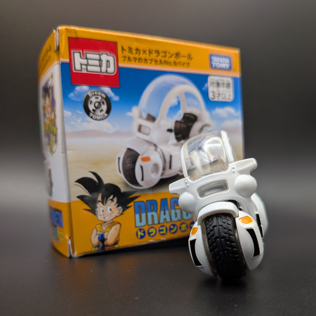 A small white Dragon Ball toy motorcycle with a clear windshield and black tire, placed in front of its orange and blue retail box featuring Dragon Ball characters and Japanese text.