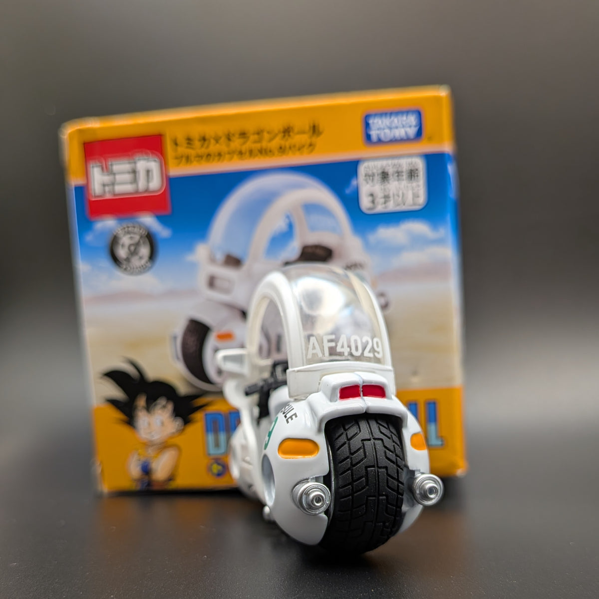 A small white and black toy motorcycle model with the number AF4029, placed in front of its original packaging box. The box features Dragon Ball Z branding and character artwork, including Goku.