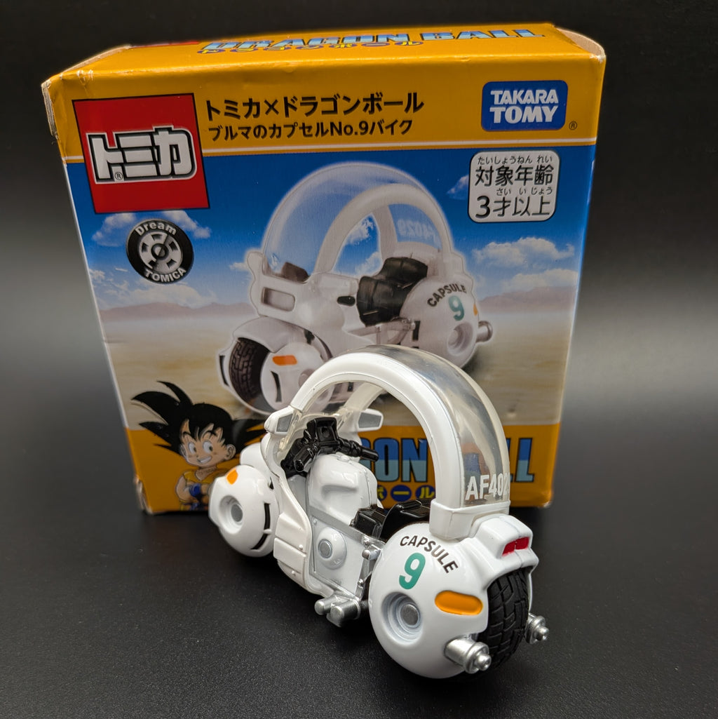 A Tomica x Dragon Ball capsule toy bike, Capsule No. 9 Bike, displayed in front of its original yellow and blue Takara Tomy packaging box on a dark gray surface.