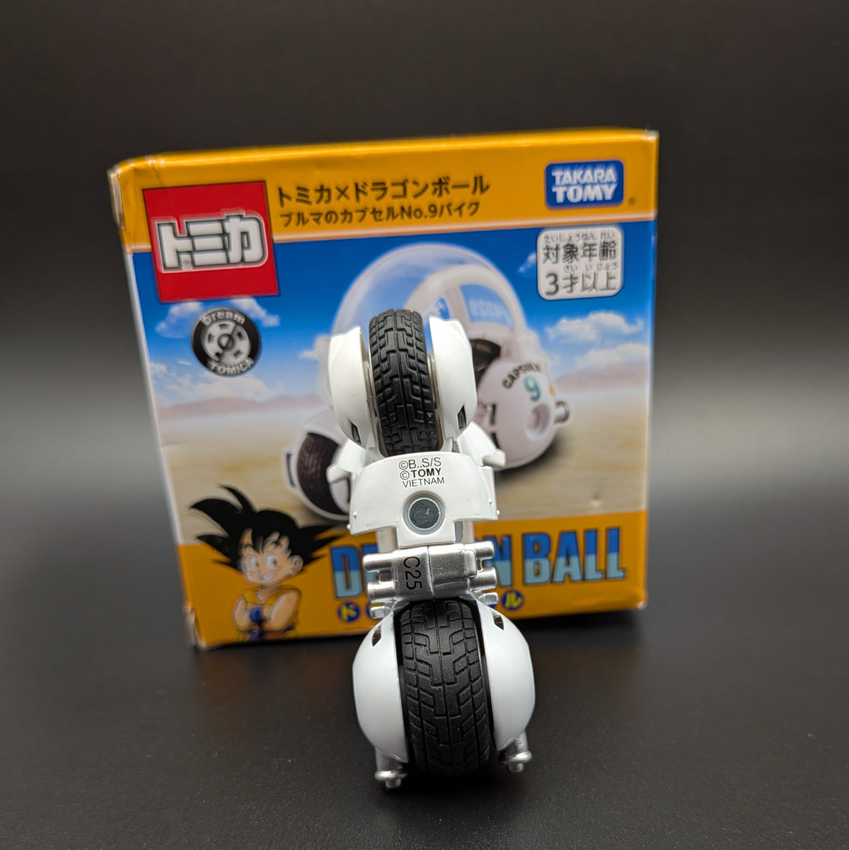 A small white and black toy motorcycle set in front of its retail box. The packaging features the Takara Tomy logo, Japanese text, and Dragon Ball character artwork on a dark gray background.