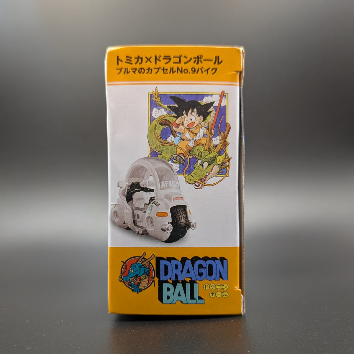 Dragon Ball x Tomica collaboration capsule toy packaging, featuring a white and yellow box with an illustration of Goku and the Dragon Ball Z vehicle Capsule on a dark gray background.