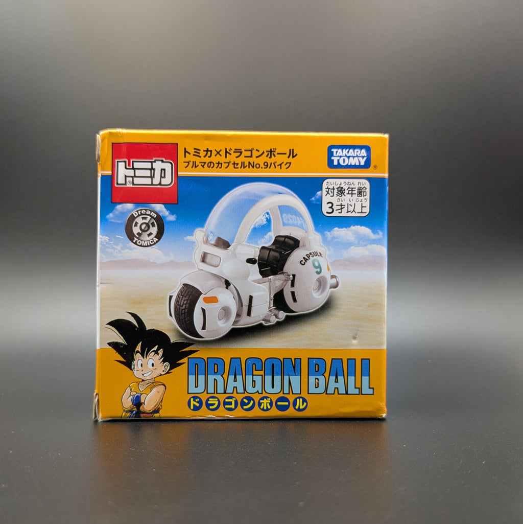 A boxed toy product labeled Dragon Ball, shown upright on a dark gray surface against a dark gray background. The package is predominantly yellow and blue with a photo of a white and black futuristic vehicle and Dragon Ball character artwork.