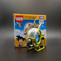 A small yellow and green toy helicopter with a clear dome cockpit stands in front of its retail box. The packaging features Dragon Ball Z artwork, including Goku, and visible brands include Tomica and Tamiya.