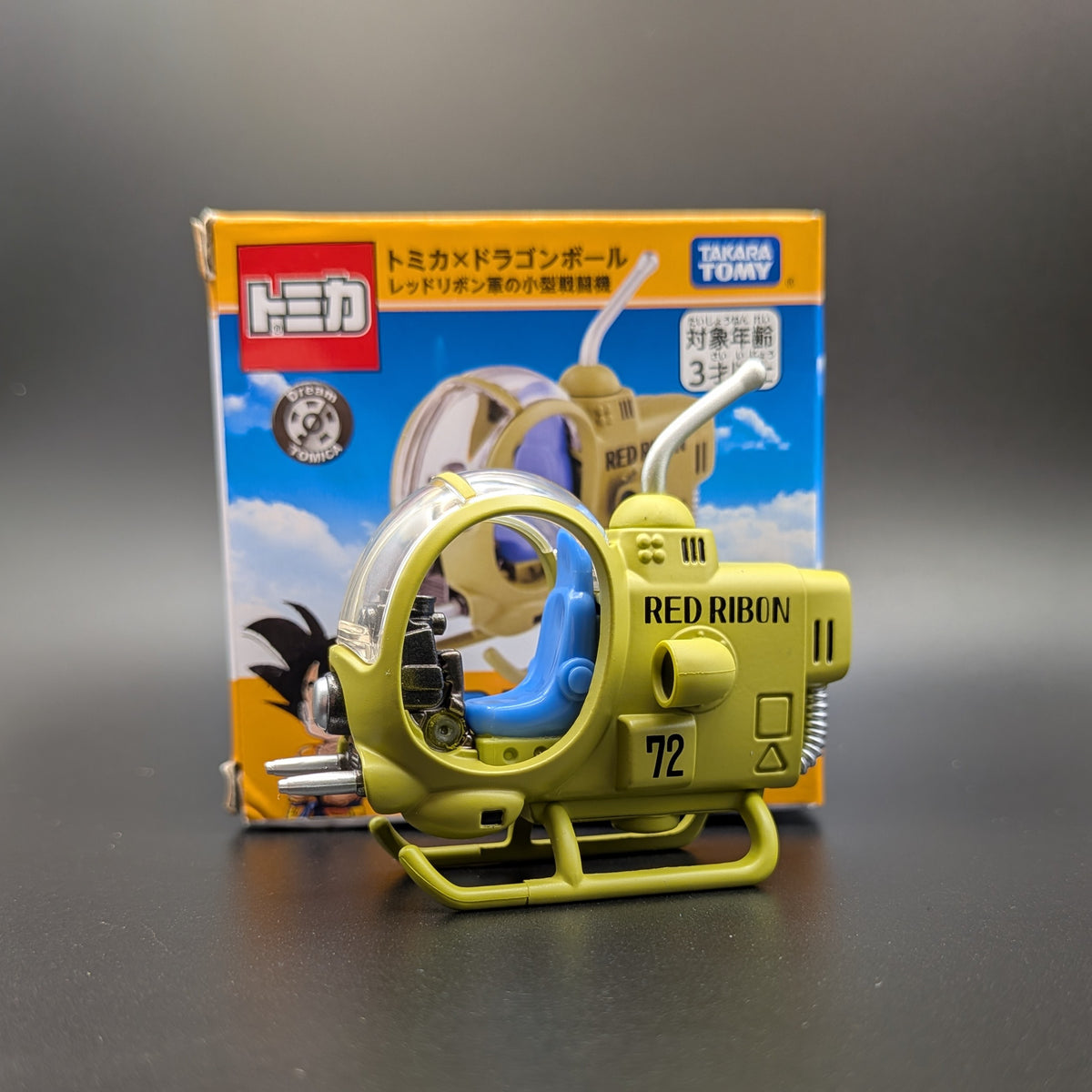 A Takara TOMY Tomica toy model, 'Red Ribbon 72', displayed on its side. In front of the packaging box on a dark gray surface.