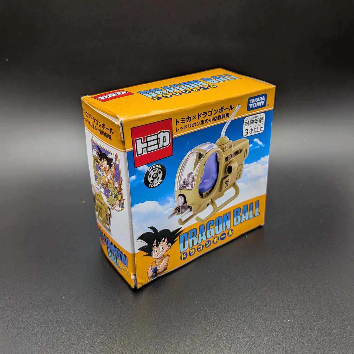 A boxed toy product labeled Dragon Ball, shown standing on a dark gray surface against a dark gray background. The box is predominantly yellow and orange with blue sky artwork and Dragon Ball character graphics.